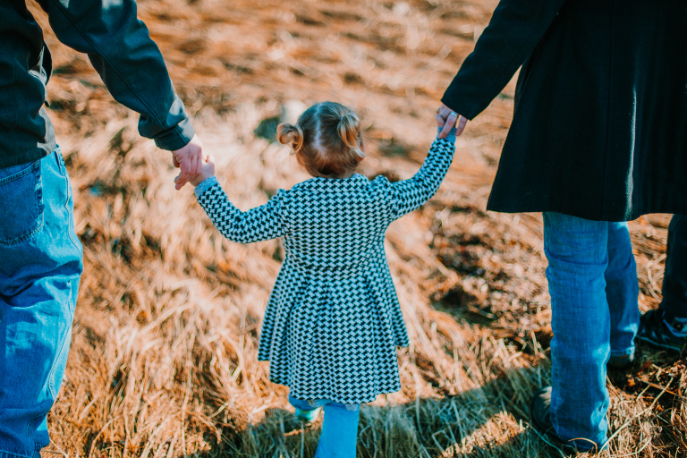 A mother and father hold the hand of their daughter in a grassy Skagway field. 