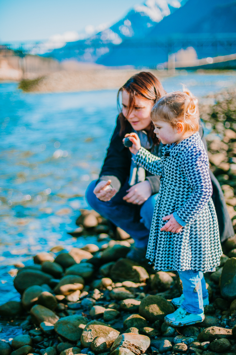 A toddler stands with her mom, ready to throw a pebble into the river. 