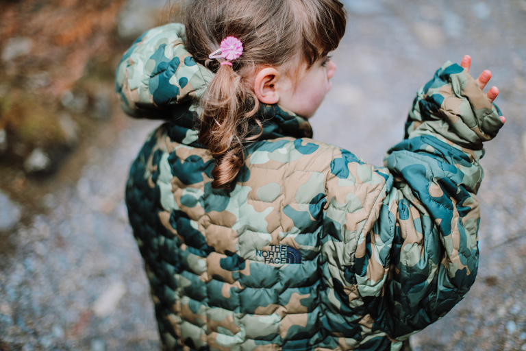 Little girl pokes her hand out of her dad's oversized camo jacket. 