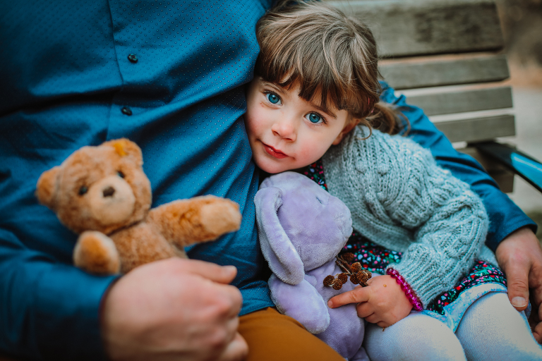 Little girl sits with her dad and her stuffed animals, holding her collected acorns and looking directly into the camera. 