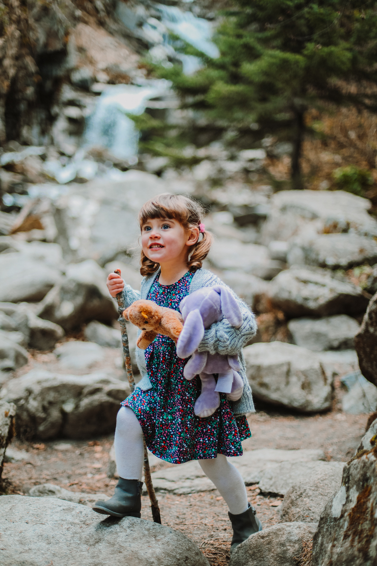 Little girl heads out on an adventure, holding onto her two stuffed animals and a walking stick. 