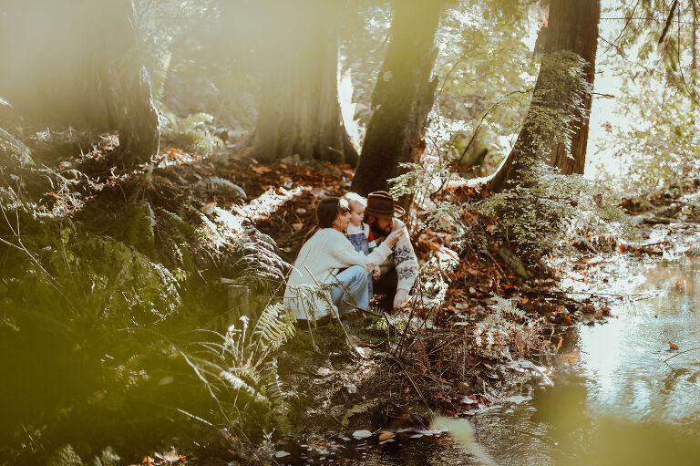 Family sits by the river in the woods, dressed in seventies attire.