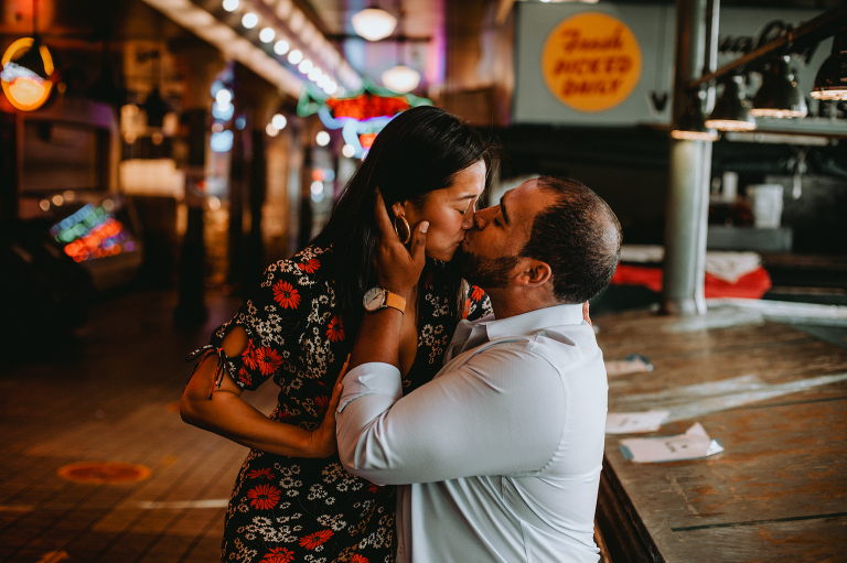 Alexis sits on Derek's lap in Pike Place Market, kissing each other on the lips.